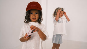 Two children wearing white shirts and bright shorts, one with a red cap, on a plain background.